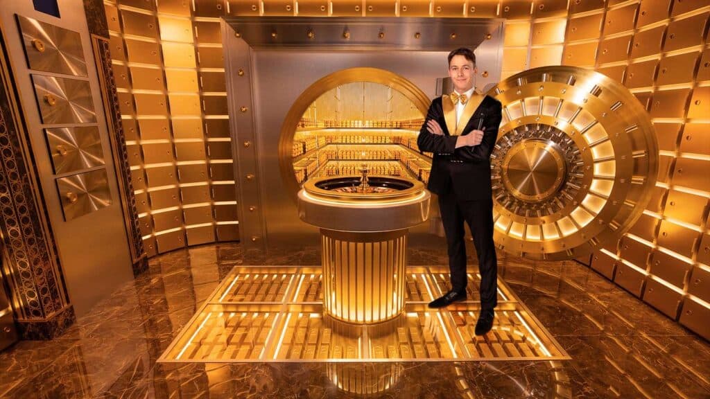 The Presenter posed in front of the roulette wheel in a golden room with a golden bank vault in the back